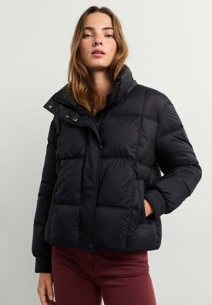Jeune femme portant une doudoune noire avec un col montant et un pantalon bordeaux, debout devant un fond uni.