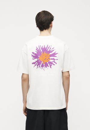 Άνδρας με κοντά σκούρα μαλλιά, φοράει λευκό T-shirt με αφηρημένο σχέδιο σε μωβ και πορτοκαλί στην πλάτη, στέκεται μπροστά σε απλή φόντο.