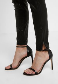 Stopy w czarnych sandałach na wysokim obcasie z paskami na kostkę oraz w czarnych spodniach z zamkami błyskawicznymi na kostkach częściowo otwartymi.