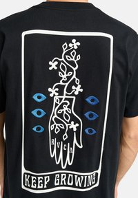 Camiseta de algodón negra con un gráfico blanco de una mano rodeada de enredaderas y flores, acentuada por ojos azules y el texto "SIGUE CRECIENDO" en la parte inferior.
