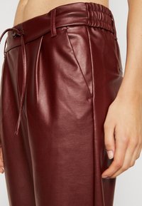 Pantalon en similicuir bordeaux avec une ceinture élastique, un cordon de serrage et des poches latérales, présentant une texture lisse et légèrement brillante.