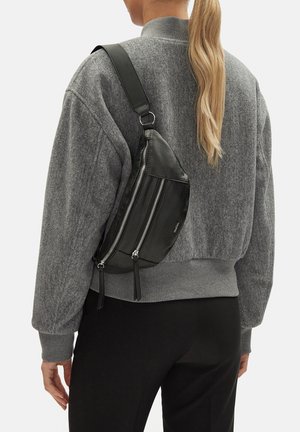 Mujer con coleta rubia, vestida con suéter gris y pantalones negros, lleva una bandolera de cuero negra con dos cremalleras verticales en la parte trasera.