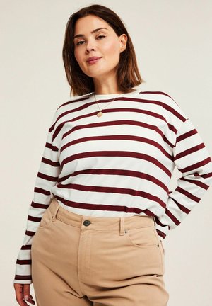 Kobieta o brązowych włosach do ramion, ubrana w białą koszulę z długim rękawem w ciemnoczerwone poziome paski oraz beżowe spodnie, pozująca z jedną ręką na biodrze.