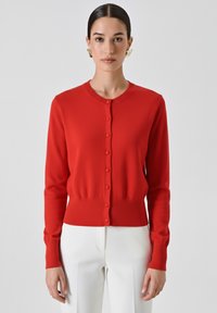 Frau mit dunklem Haar in einem glatten Dutt, die goldene Ohrringe, eine rote Knopfpulloverjacke und weiße Hose trägt und vor einem schlichten Hintergrund steht.