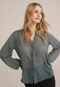 Bluză transparentă verde cu un model geometric, închidere cu nasturi și mâneci lungi. Material texturat cu un design ușor și fluid.