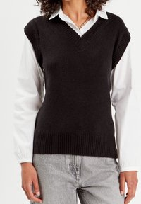 Veste noire en maille avec un col en V, ourlet côtelé et manches courtes, portée sur une chemise blanche. Texture douce, coupe ajustée.