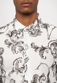 Camisa blanca con patrón floral en negro y gris. Presenta un cuello clásico y cierre frontal con botones y herrajes visibles. Textura suave.