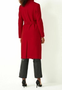 Manteau en mélange de laine rouge avec une ceinture, manches longues et fente au dos. Assorti à un pantalon en cuir noir et des escarpins noirs à clous.