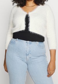 Cárdigan corto blanco y esponjoso con una capa interior negra, que presenta mangas largas y un escote redondeado, combinado con jeans de tiro alto en azul claro.