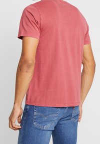 T-shirt z krótkim rękawem i okrągłym dekoltem w kolorze wyblakłego różu, wykonany z bawełny. Charakteryzuje się luźnym krojem i przeszywanym dołem. Noszony z niebieskimi dżinsami.