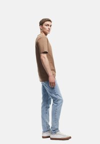 Camiseta de algodón marrón de manga corta, combinada con jeans de mezclilla azul claro. El modelo lleva zapatillas blancas con suela marrón, posando de perfil.
