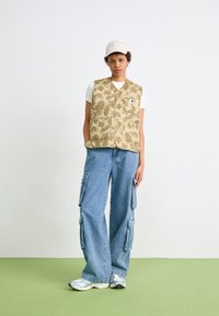 Tan camo puffer vest over a white t-shirt, paired with light blue wide-leg cargo jeans. Includes pockets and a beige cap.