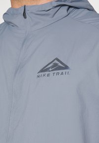 Vaaleansininen Nike Trail -takki, jossa on vetoketjullinen etuosa, musta logo ja geometrinen kuvio rinnassa, valmistettu sileästä teknisestä kankaasta.