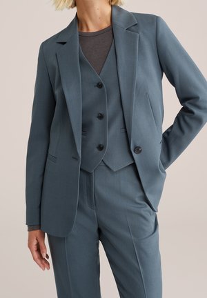Kvinde iført en blå tredelt dragt med blazer, knappet vest og bukser, kombineret med en mørkegrå langærmet top under.