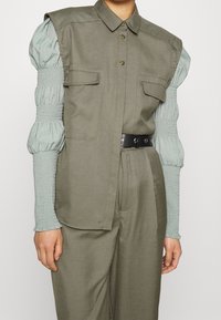 Koszula w kolorze oliwkowym o strukturalnym fasonie, z przednimi kieszeniami i czarnym paskiem. Miękki, marszczony materiał rękawów w kolorze miętowym dodaje kontrastu.