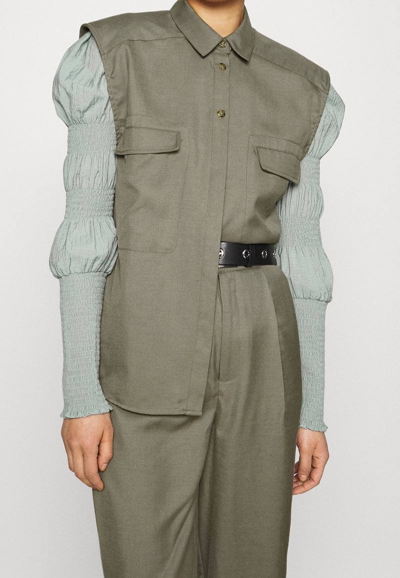 Koszula w kolorze oliwkowym o strukturalnym fasonie, z przednimi kieszeniami i czarnym paskiem. Miękki, marszczony materiał rękawów w kolorze miętowym dodaje kontrastu.