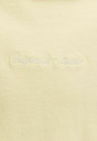 Tissu jaune pâle avec un texte brodé blanc subtil indiquant « Originals Studio » en police cursive.