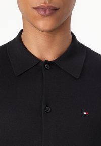 Čierne polo tričko vyrobené z pleteného materiálu, s klasickým golierom, dvoma gombíkmi a malým logom v červenej, bielej a modrej farbe na hrudi.