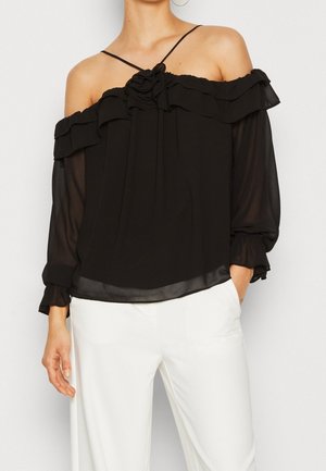 Kvinde iført en sort off-shoulder bluse med gennemsigtige lange ærmer, flæsedetalje og tynde stropper, kombineret med hvide bukser med høj talje.