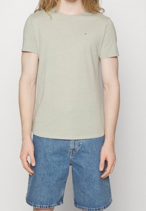 Personne portant un t-shirt vert clair à manches courtes avec un petit logo sur la poitrine et un short en jean bleu, debout devant un fond uni.