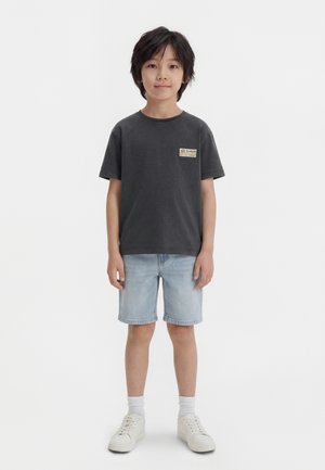 Chłopiec stoi zwrócony twarzą do przodu, ubrany w ciemnoszary t-shirt, jasne dżinsowe spodenki, białe skarpetki i białe trampki na białym tle.