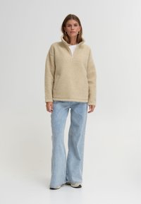 Beiger Fleece-Pullover mit einem Halb-Reißverschluss-Kragen, kombiniert mit hellblauen ausgestellten Jeans und schwarzem Schuhwerk. Glatte Textur, lockere Passform.