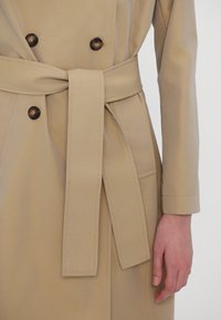 Trench-coat beige avec des boutons croisés, une ceinture en tissu nouée à la taille et la main d'une personne reposant à côté du manteau.