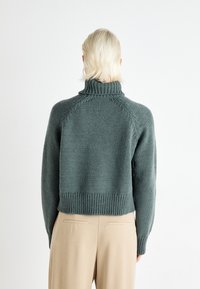 Knitted green turtleneck sweater with ribbed cuffs and hem, featuring a relaxed fit and textured fabric. Paired with beige trousers.