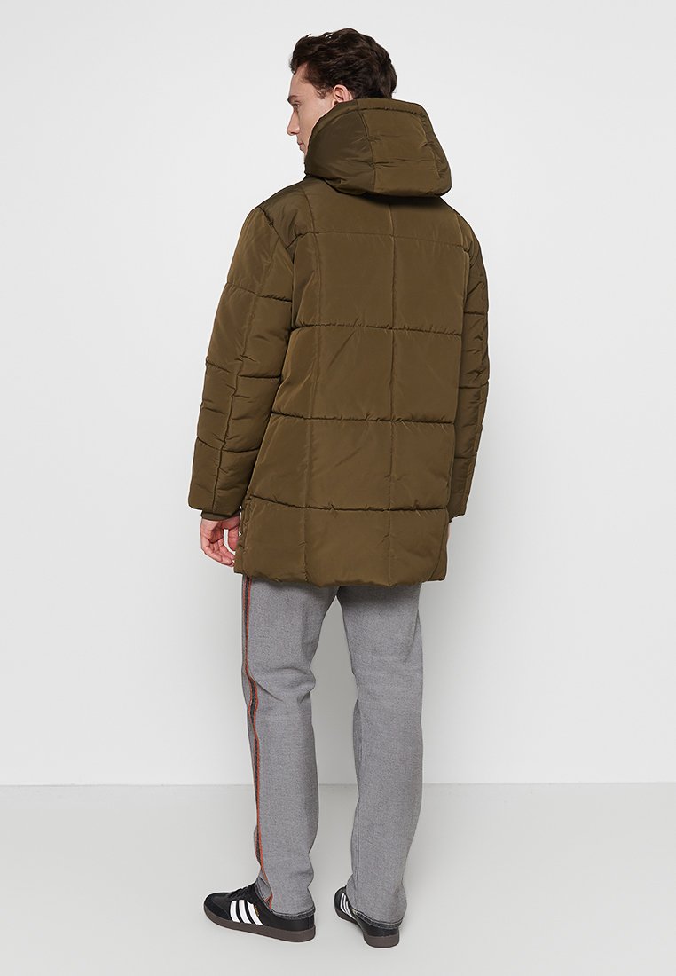 Olivenfarbene gepolsterte Jacke mit Kapuze, die ein gesteppter Entwurf und einen längeren Schnitt aufweist. Kombiniert mit grauen Hosen und schwarzen Schuhen.