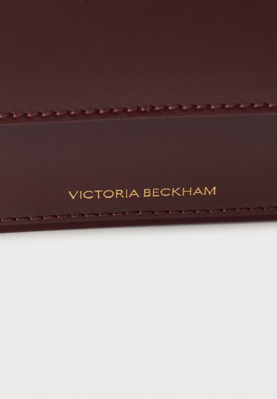 Κοντινή λήψη αντικειμένου από δέρμα μπορντό με τακτοποιημένα ραφές και χρυσή ανάγλυφη επιγραφή "Victoria Beckham".