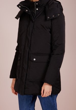 Zwart gewatteerd jack met hoge kraag en klepzakken met drukknopen, gedragen door een persoon in blauwe jeans, zichtbaar vanaf de schouders tot de heupen.