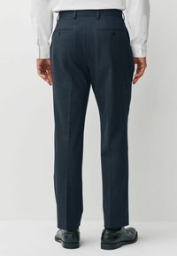 Dark blue tailored trousers with a textured fabric, slim fit, flat front, and two back pockets with button closures. Paired with black shoes.