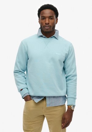 Hombre adulto con cabello rizado oscuro que lleva una sudadera azul claro sobre una camisa a rayas azul, pantalones beige y un reloj de pulsera dorado, de pie con las manos en los bolsillos.