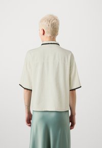 Cămașă scurtă bej deschis, cu mânecă scurtă și un guler negru cu margini ondulate. Materialul pare texturat, cu o croială lejeră și un design minimalist.