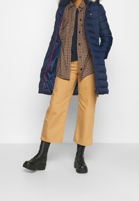 Donkerblauwe pufferjas met capuchon, geruit bruin shirt, donkere broeken en zwarte veterschoenen. Bevat een ritssluiting en contrasterende rode details.