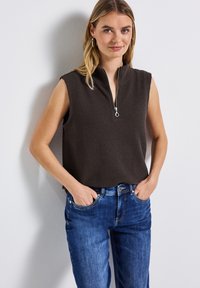 Mörkbrun ärmlös topp med en texturerad, våffelmönstrad yta och en halvmatchande dragkedja vid halslinjen. Kombinerad med blå denimjeans där fickorna är synliga.