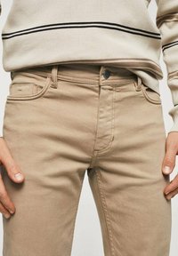 Pantalones beige con bolsillos frontales y botón, combinados con un suéter beige con rayas horizontales negras, llevados por una persona.