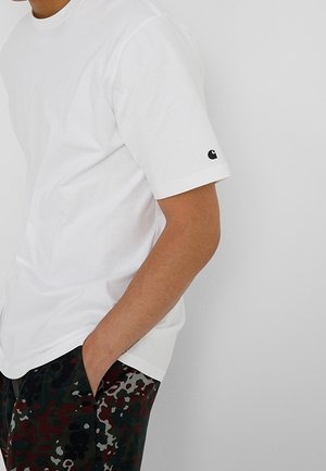 Hombre vistiendo una camiseta blanca lisa de manga corta con un pequeño logo en la manga y pantalones de camuflaje multicolor oscuros con una mano en el bolsillo.