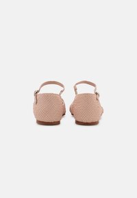 Paire de sandales plates tissées beige avec brides à la cheville et petites boucles, vues de dos sur un fond uni.