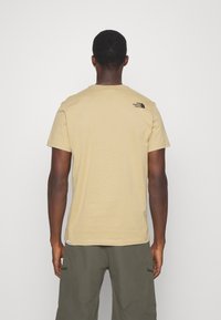 Muž v béžovém tričku s krátkým rukávem se malým černým logem "The North Face" na horní části zad, v olivově zelených kalhotách, otočený čelem k nám.