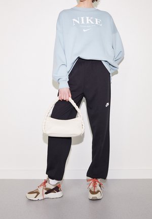 Person iført lyseblå Nike sweatshirt, sorte Nike bukser, beige og brune sneakers med røde snørebånd, der holder en hvid håndtaske med flettede håndtag.