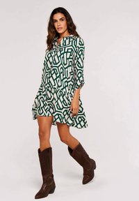 Weißes Kleid mit grünem geometrischem Muster, langen Ärmeln und volantsaum. Kombiniert mit braunen kniehohen Stiefeln.