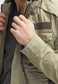 Light olive green jacket made of woven fabric, featuring a full zipper, snap buttons, and a chest pocket with a textured accent.