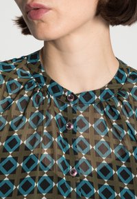 Mönstrad blus med geometrisk diamantdesign i blå, svart och grön nyanser. Har en rynkad halsringning och två runda knappar.