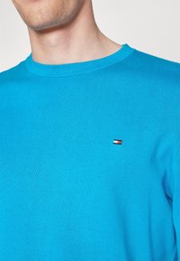 Pull en tricot bleu clair avec col rond côtelé, présentant un petit logo rouge, blanc et bleu sur le côté gauche de la poitrine. Design simple, tissu texturé.