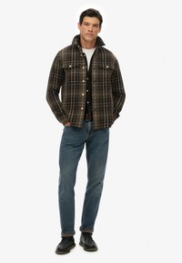 Camisa de franela a cuadros en marrón, negro y amarillo, con dos bolsillos en el pecho y un cuello de solapa, combinada con vaqueros azules y zapatos negros.