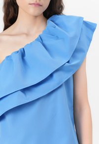 Haut bleu clair avec un volan asymétrique unique sur l'épaule. Tissu doux avec une texture lisse, présentant une coupe décontractée et des lignes épurées.