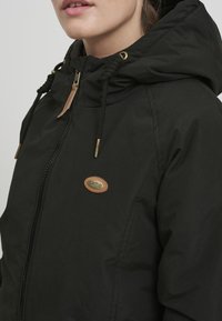 Giacca nera con cappuccio, dotata di zip, chiusura con cordino e una toppa in pelle con logo. Il materiale appare liscio con una finitura leggermente opaca.