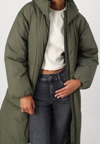 Abrigo acolchado largo en verde oliva con capucha, que presenta secciones acolchadas y una cremallera frontal. Combinado con una camiseta corta blanca y jeans grises.