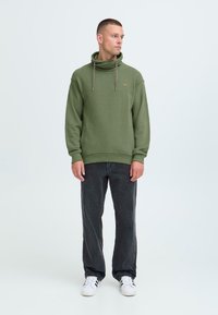 Felpa verde con cappuccio e cordini, polsini a coste e dettaglio con logo. Abbinata a jeans neri e sneakers bianche.